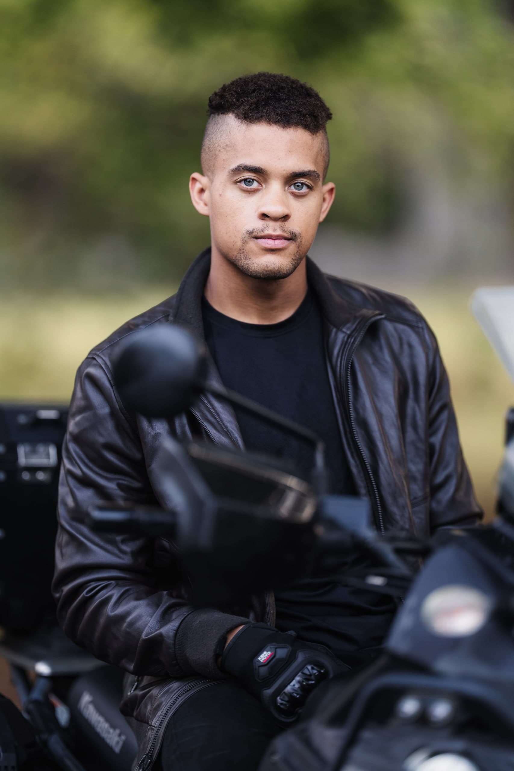 A young man in a leather jacket portrait, motorcycle beside him.