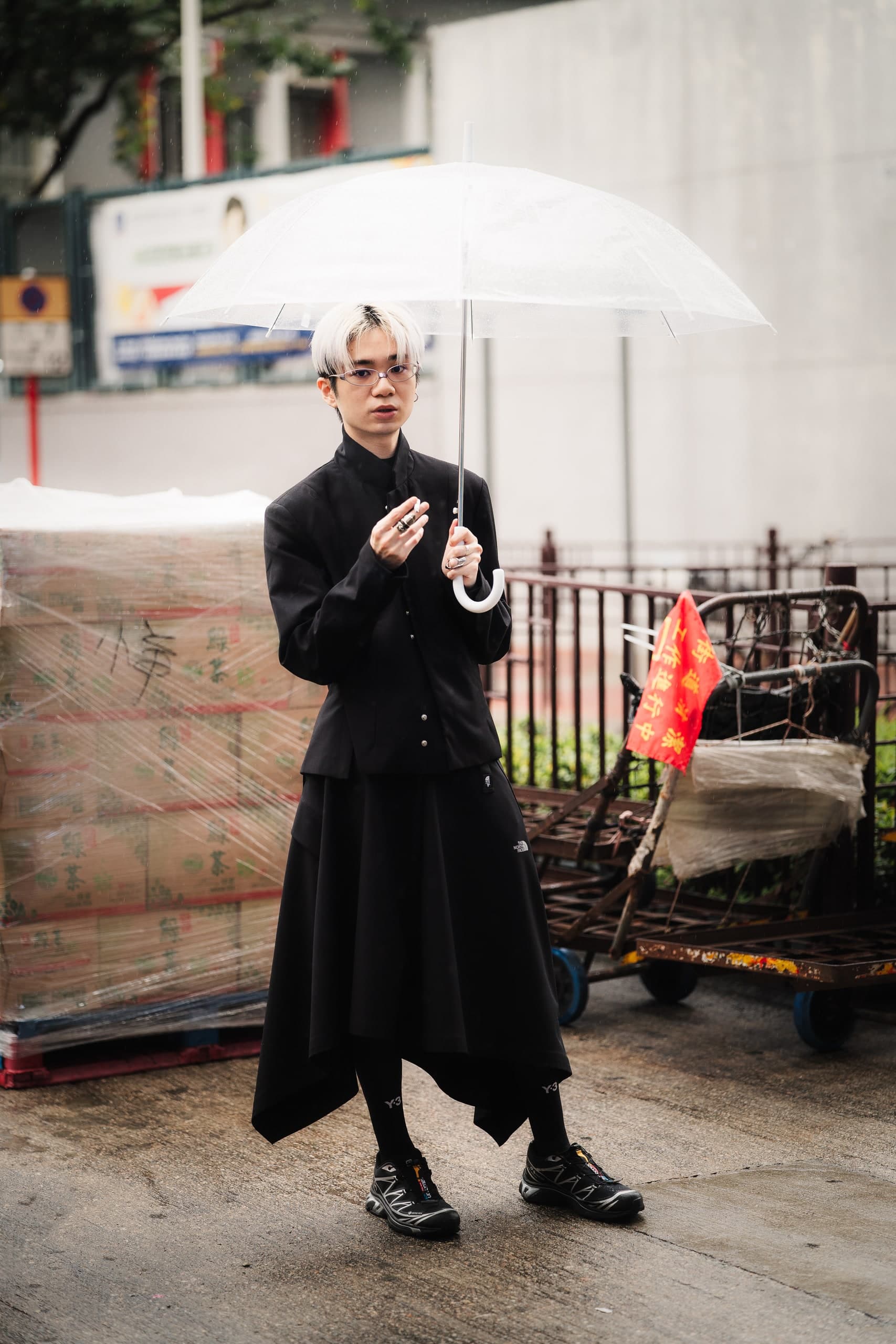 A young man in dark, draped clothing holding a clear umbrella on a city street.