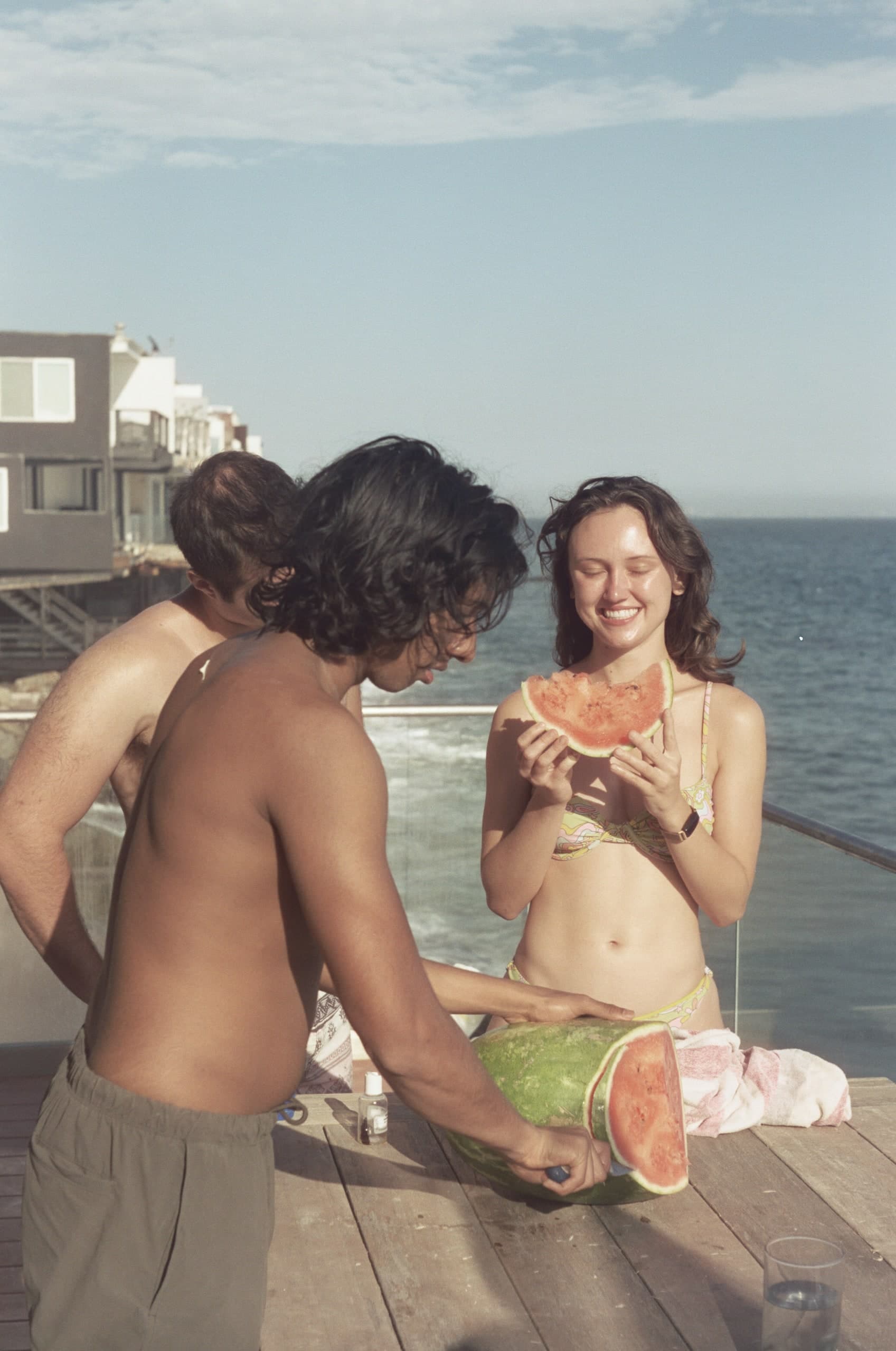 Friends sharing watermelon on a dock, a beach house behind them, film photograph.
