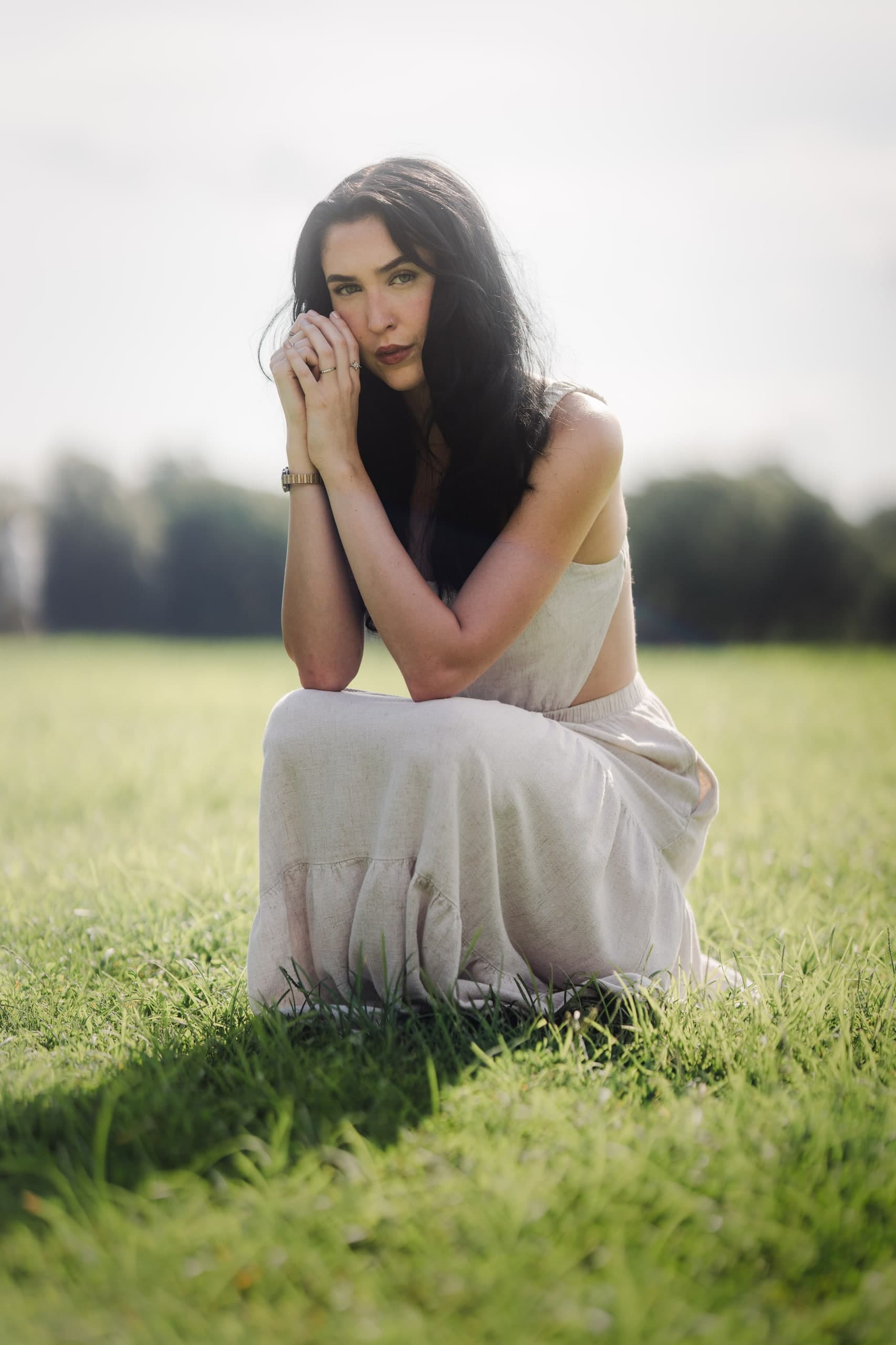 A woman in a cream dress kneeling in a sunlit field, one hand resting thoughtfully under her chin.