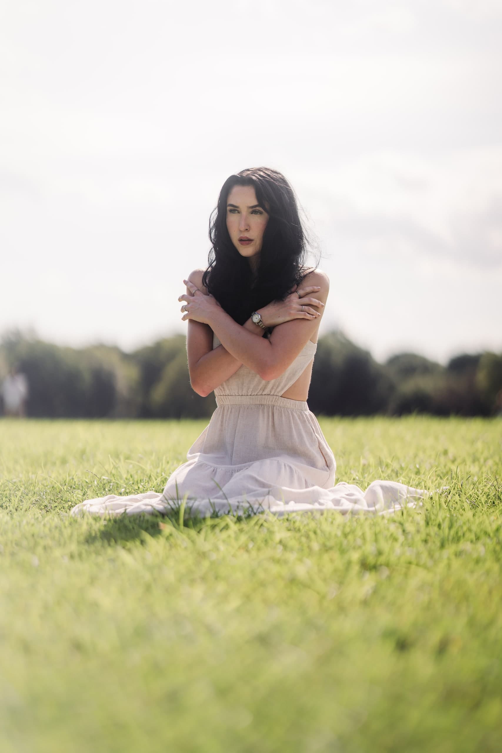 A woman in a cream dress sitting in a grass field, arms wrapped around herself, looking into the distance.